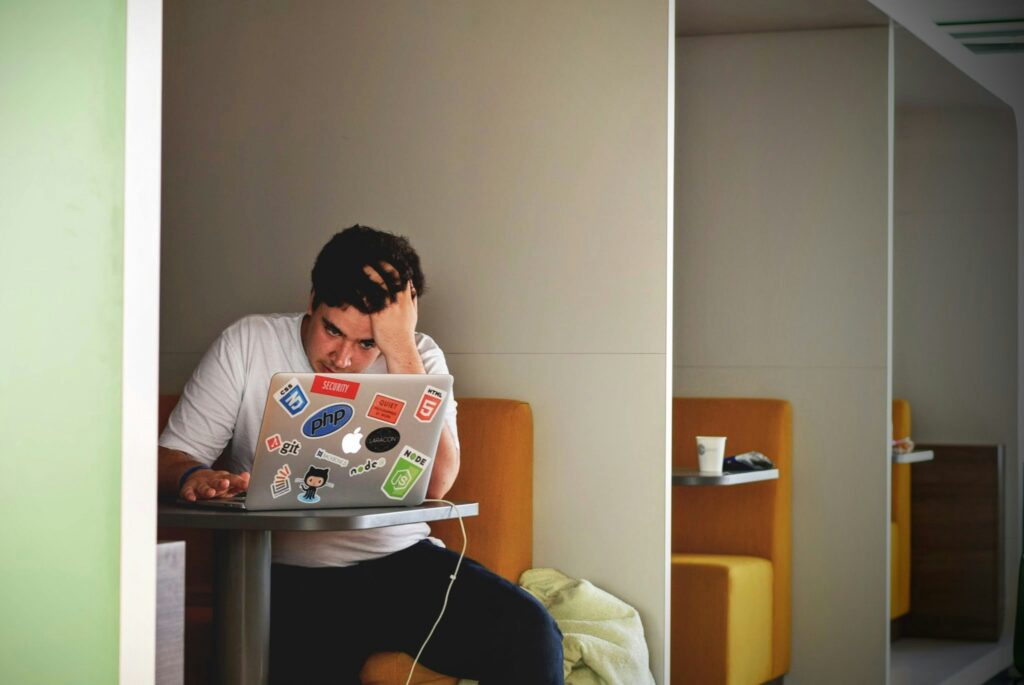 jovem de camiseta branca e mão na cabeça sentado num banco, olhando para o notebook repleto de adesivos que está numa mesa à sua frente.